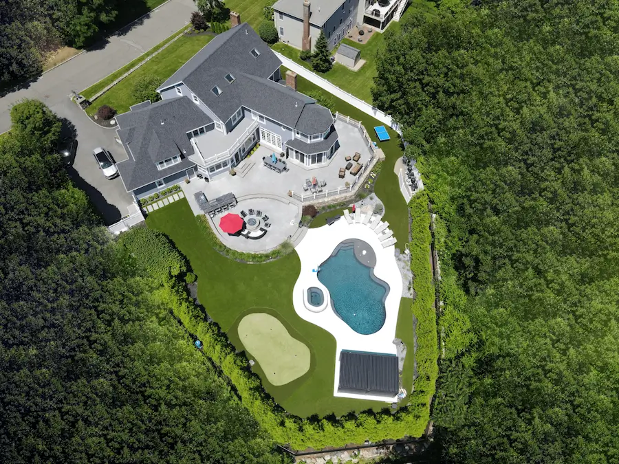 aerial view luxury landscaping in Lynnfield, Ma. Stone masonry around a blue pool and custom golf putting green sit on the expansive manicured lawn which is lined with evergreen shrubs.